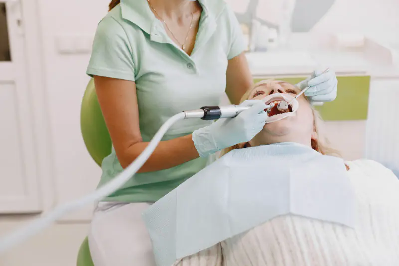 Praticienne en tunique vert d’eau nettoyant les dents d’une patiente allongée au fauteuil, lors d’une séance de soins de prévention