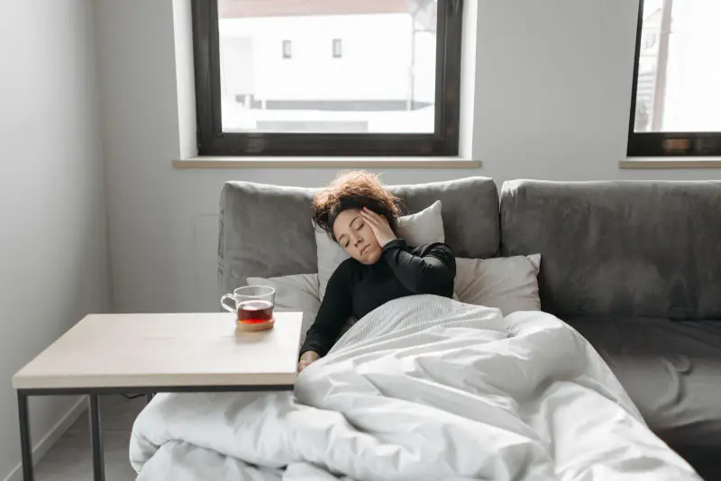 Femme allongée sur un canapé sous une couette, la main posée sur la joue, avec une boisson sur la table basse à côté d’elle
