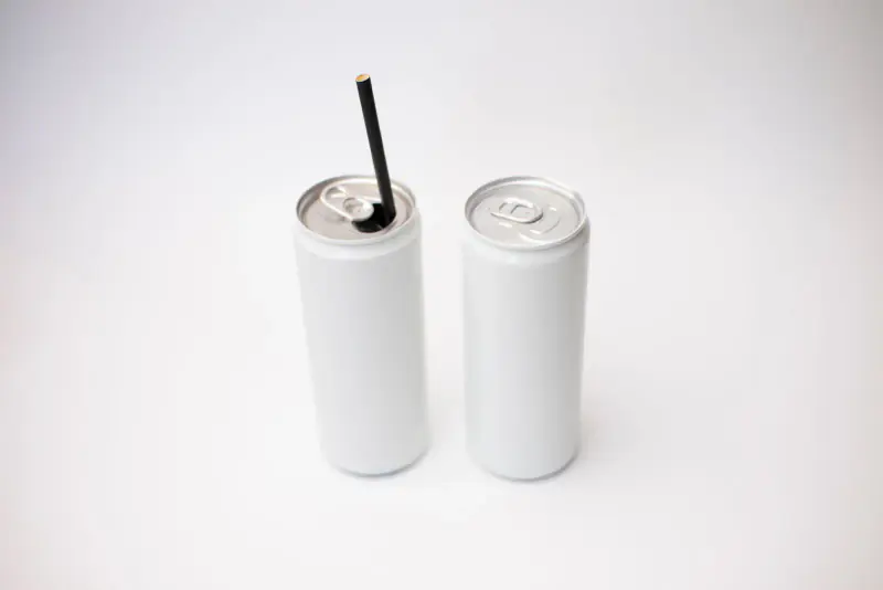 Deux canettes fines, blanches et sans marque, dont une ouverte avec une paille noire, posées sur un fond clair et lumineux