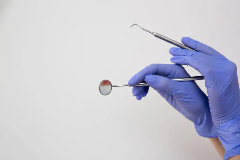 Mains gantées tenant un miroir buccal et une sonde dentaire, les deux instruments de base de l’examen des caries, sur fond blanc