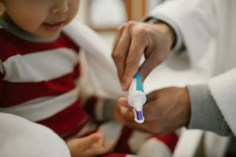 Mains d’un adulte déposant du dentifrice sur la petite brosse à dents d’un jeune enfant vêtu d’un haut rayé rouge et blanc