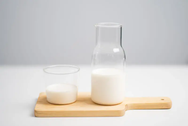 Petit verre de lait et carafe de lait posés sur une planche en bois, sur une table blanche baignée d’une lumière douce