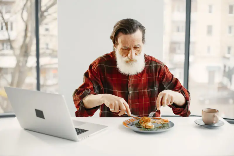 Homme âgé attablé devant une assiette dans une pièce lumineuse, coupant son repas en petits morceaux avec couteau et fourchette