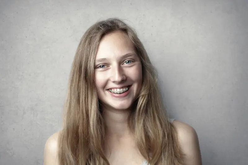 Jeune femme souriante portant des bagues orthodontiques, photographiée de face devant un mur gris clair, en lumière douce