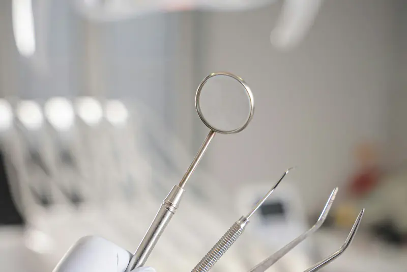 Main gantée tenant un miroir, une sonde et des précelles d’examen dentaire, devant un fauteuil de soins flou