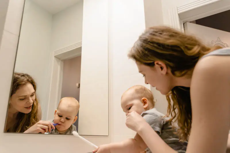 Jeune femme brossant les dents d’un bébé avec une petite brosse violette, scène vue dans le miroir d’une salle de bains claire