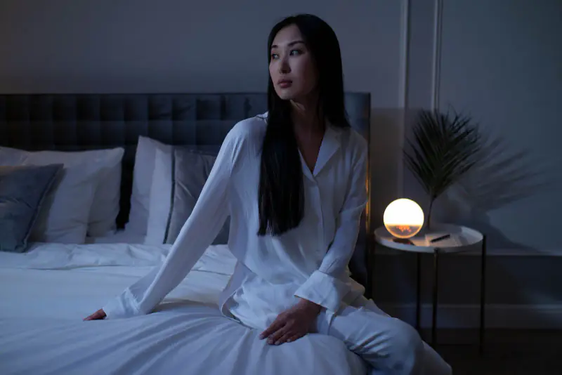 Femme en pyjama blanc réveillée en pleine nuit, assise dans son lit près d’une lampe de chevet allumée, le regard ailleurs
