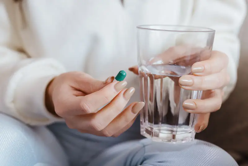 Mains d'une personne en pull clair tenant une gélule entre deux doigts et un verre d'eau
