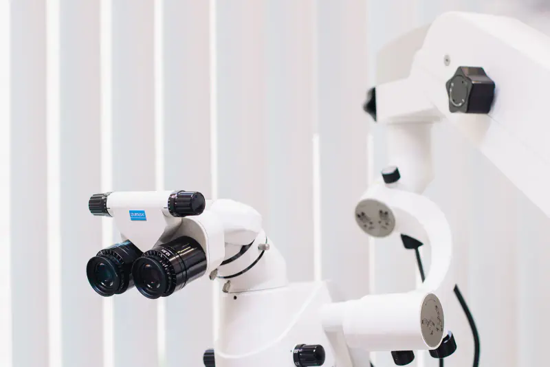 Microscope opératoire dentaire blanc monté sur son bras articulé, devant des stores clairs dans une salle de soins