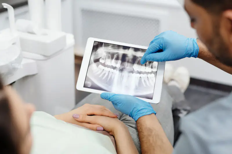Mains gantées montrant une radiographie panoramique des deux mâchoires sur une tablette à une patiente installée au fauteuil