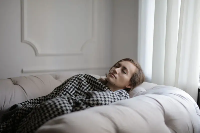 Femme qui se repose, les yeux fermés, allongée sous un plaid sur un canapé clair, la tête surélevée, dans une lumière douce