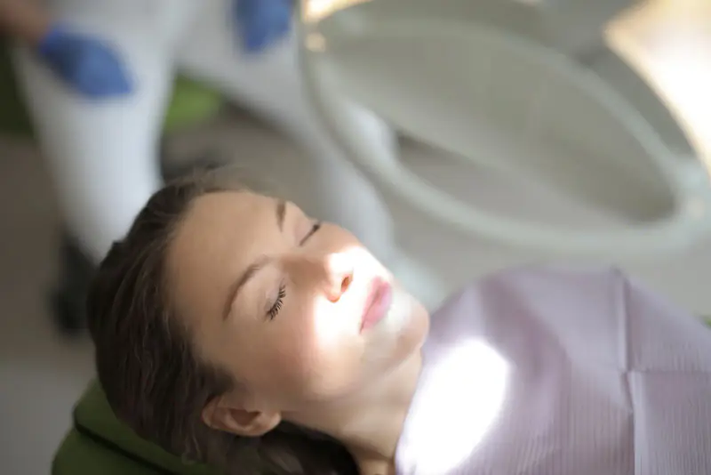 Relaxed patient with her eyes closed, reclining in a green dental chair under the soft glow of the operating light