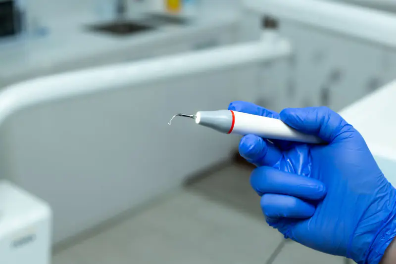 Main gantée tenant un détartreur à ultrasons à embout éclairé, l’instrument qui sert à enlever le tartre, dans un cabinet clair