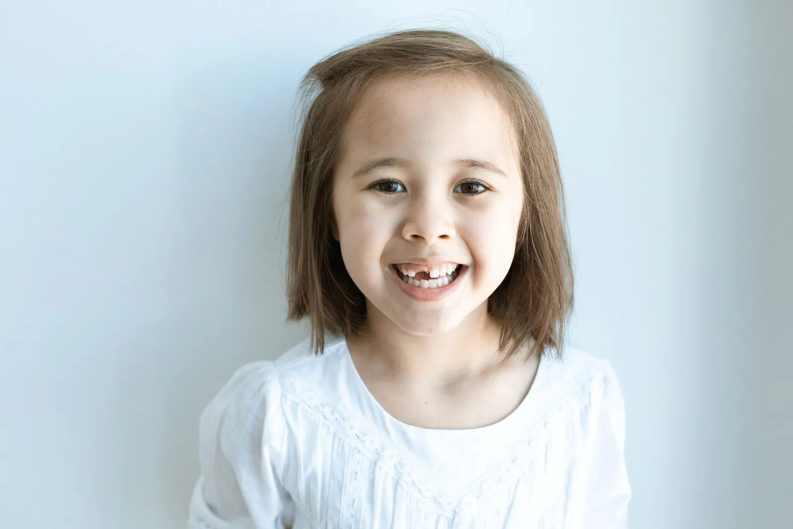 Petite fille souriante en haut blanc, à qui il manque une dent de devant, photographiée sur un fond clair et lumineux