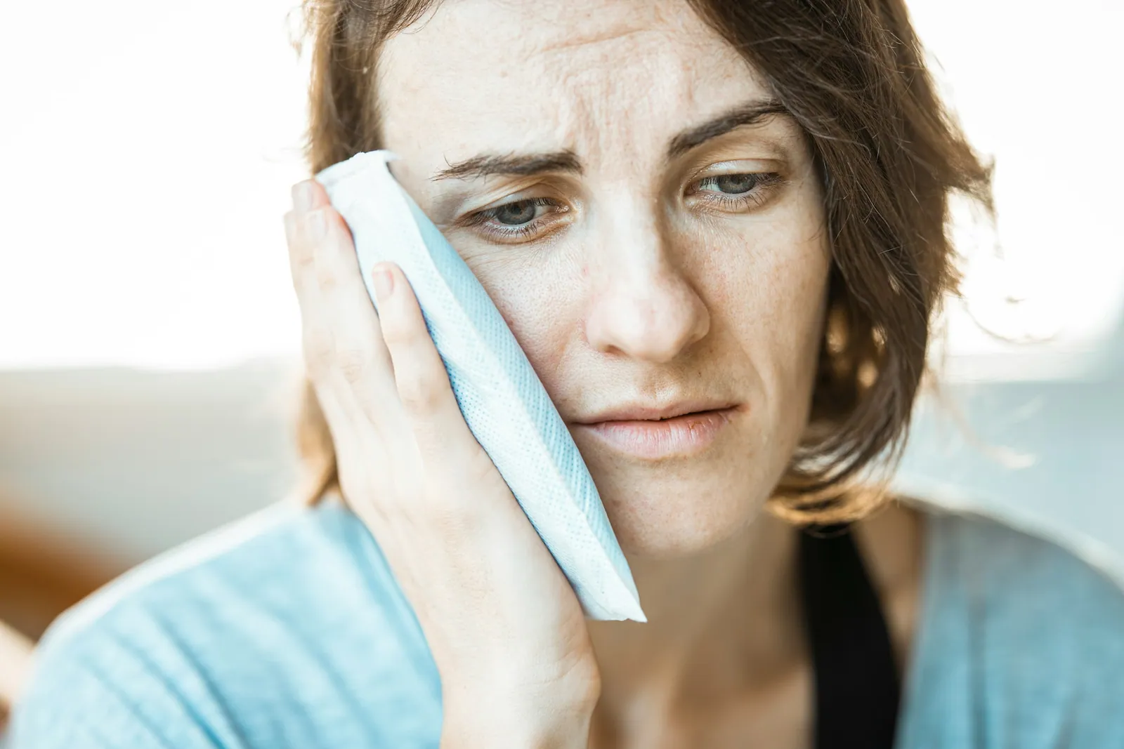 Femme appliquant contre sa joue une poche de froid enveloppée dans un tissu, l’air gêné, dans une lumière douce de jour