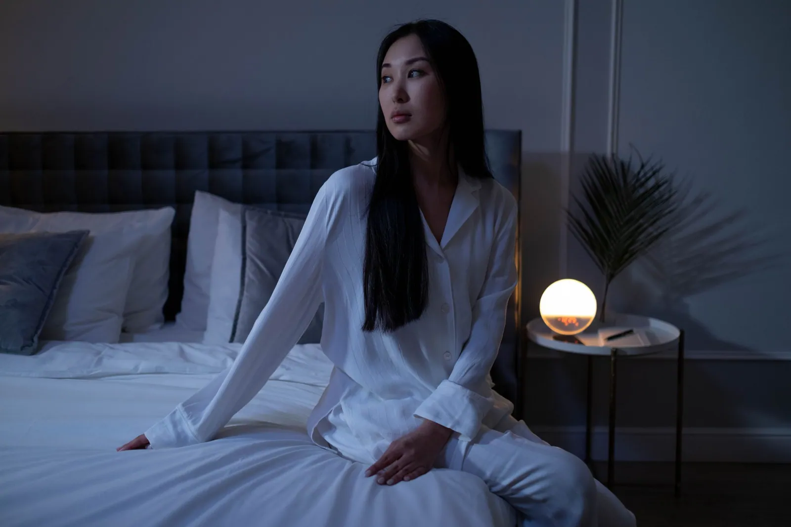Femme en pyjama blanc réveillée en pleine nuit, assise dans son lit près d’une lampe de chevet allumée, le regard ailleurs