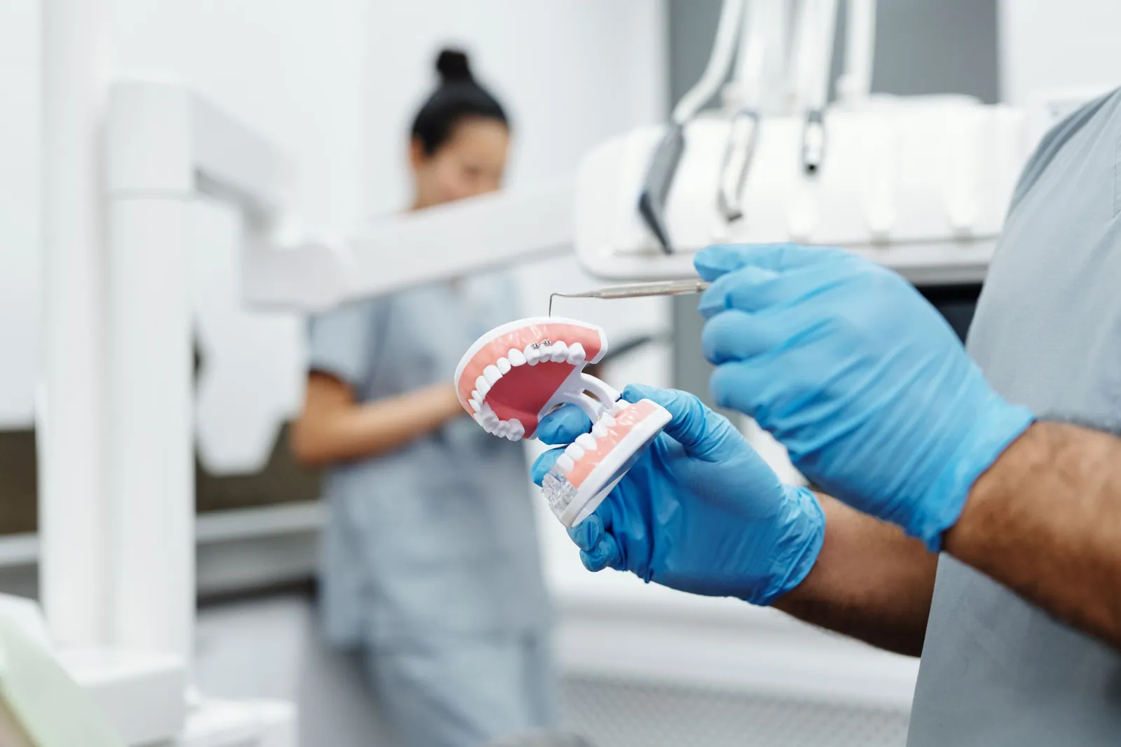 Gloved hands pointing a dental instrument at the gum of a jaw model, in a bright, uncluttered surgery