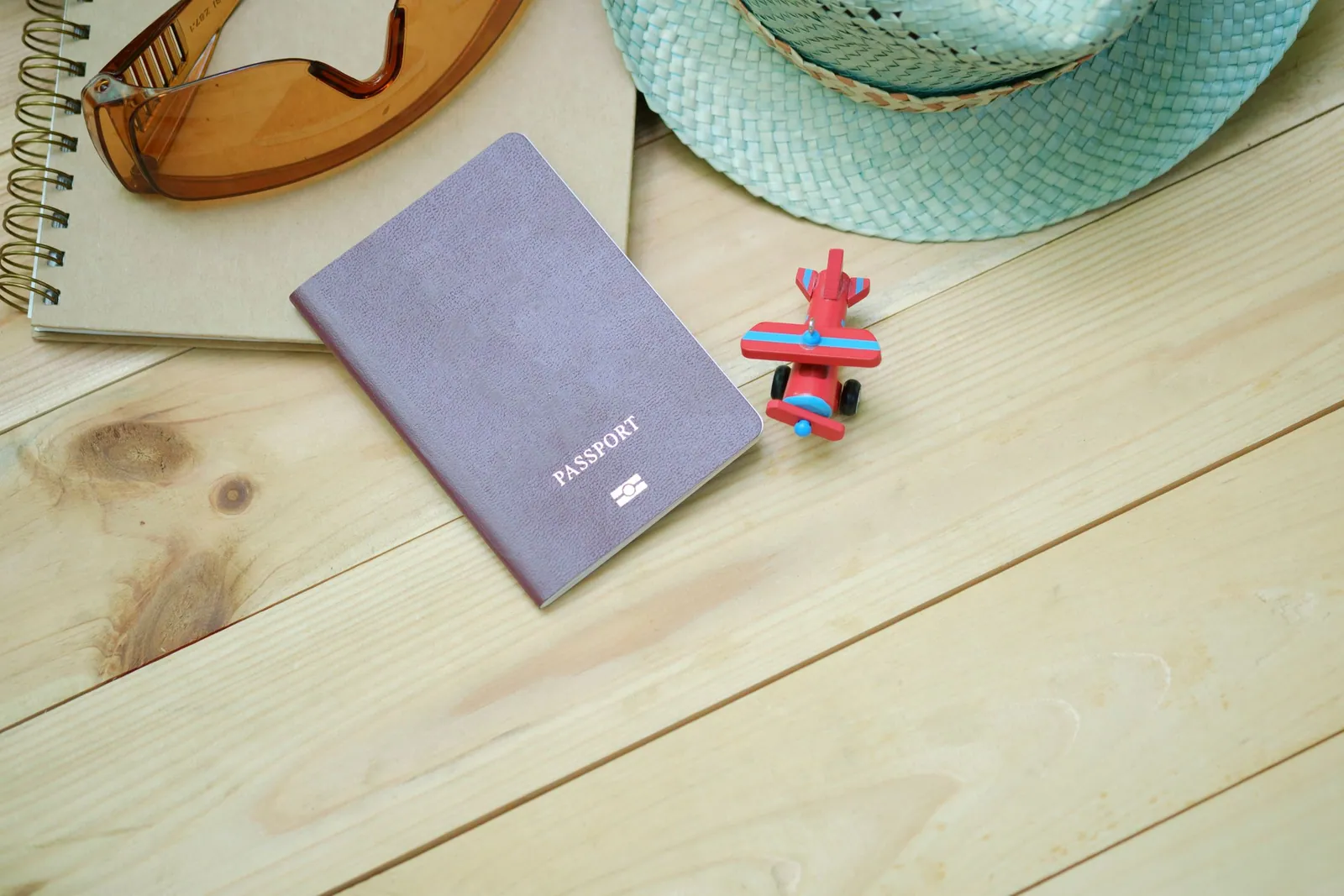 Passeport, carnet, lunettes de soleil, chapeau de paille vert d’eau et petit avion jouet sur une table en bois clair