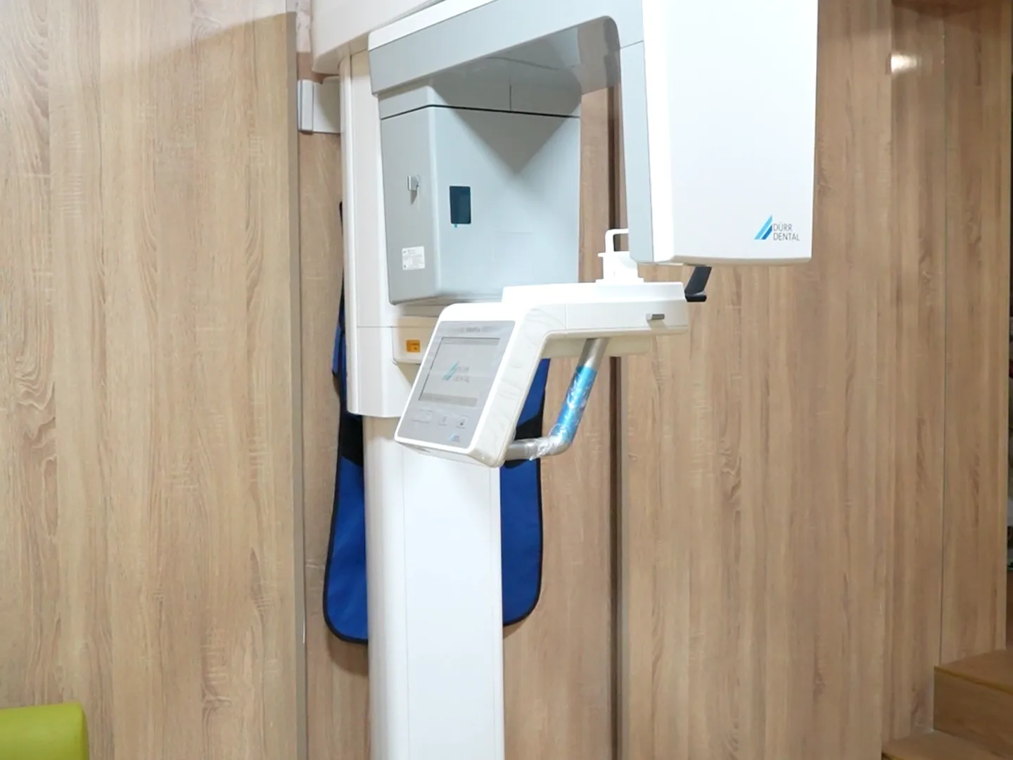 Das Cone-Beam-Gerät der Praxis, ein weißes, an der Wand montiertes Gerät in einem Behandlungsraum: oben der drehbare C-Bogen und das Aufbissstück, an dem sich die Patientin oder der Patient positioniert, ein Touchscreen am waagerechten Positionierarm, der nach vorne ragt, etwa auf Brusthöhe und der Schriftzug Dürr Dental auf dem Arm. Dahinter eine mit Holzpaneelen verkleidete Wand und eine blaue Schürze, die hinter diesem Arm am Gerät hängt; unten links die Ecke eines kleinen blassgrünen Hockers auf Holzboden.