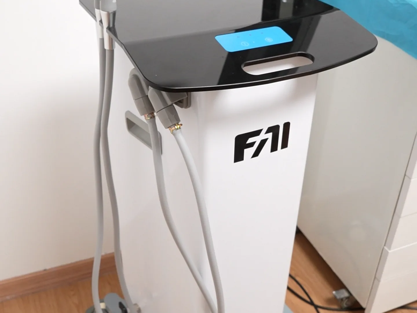 Eine weiße Gerätesäule mit schwarzer Glasplatte, einem kleinen blau leuchtenden Display in der Oberseite und zwei dicken grauen Schläuchen, die seitlich eingehängt sind; sie steht auf einem Holzboden vor einer schlichten Wand.